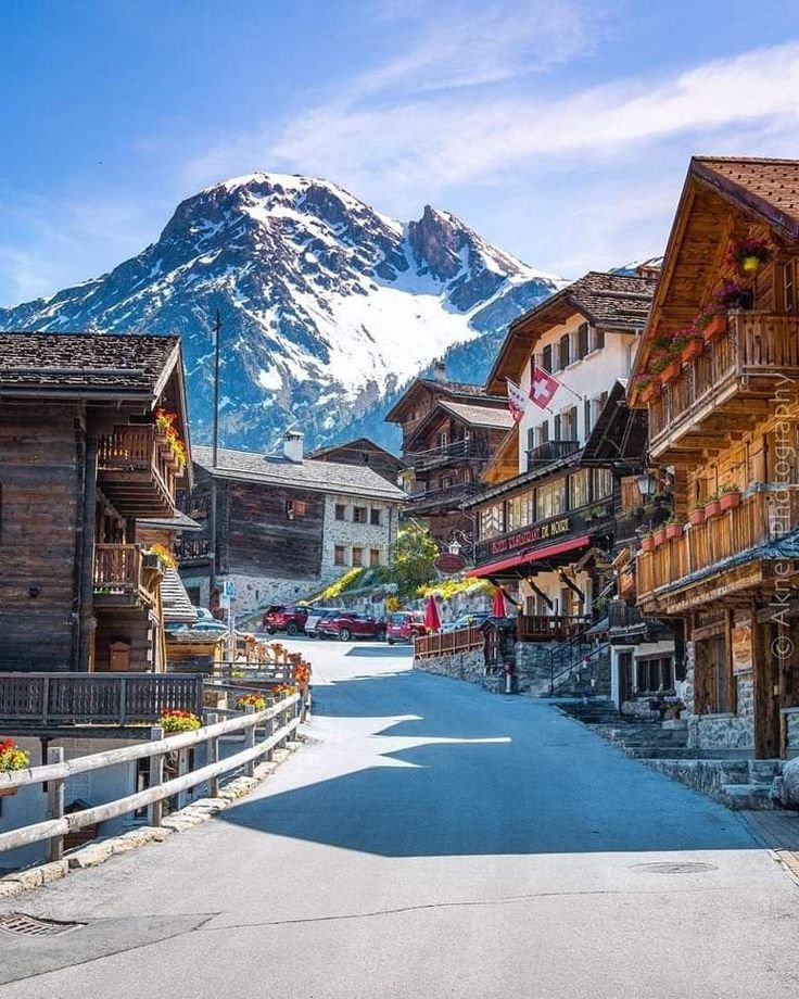 This may contain: an empty street with wooden buildings and mountains in the backgrouds, surrounded by snow - capped mountains