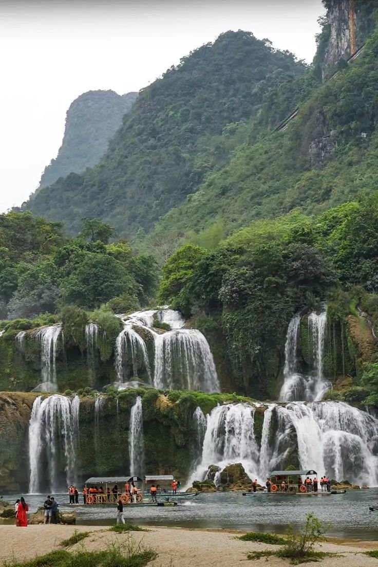 This may contain: people standing in front of waterfalls on the side of a mountain with green trees