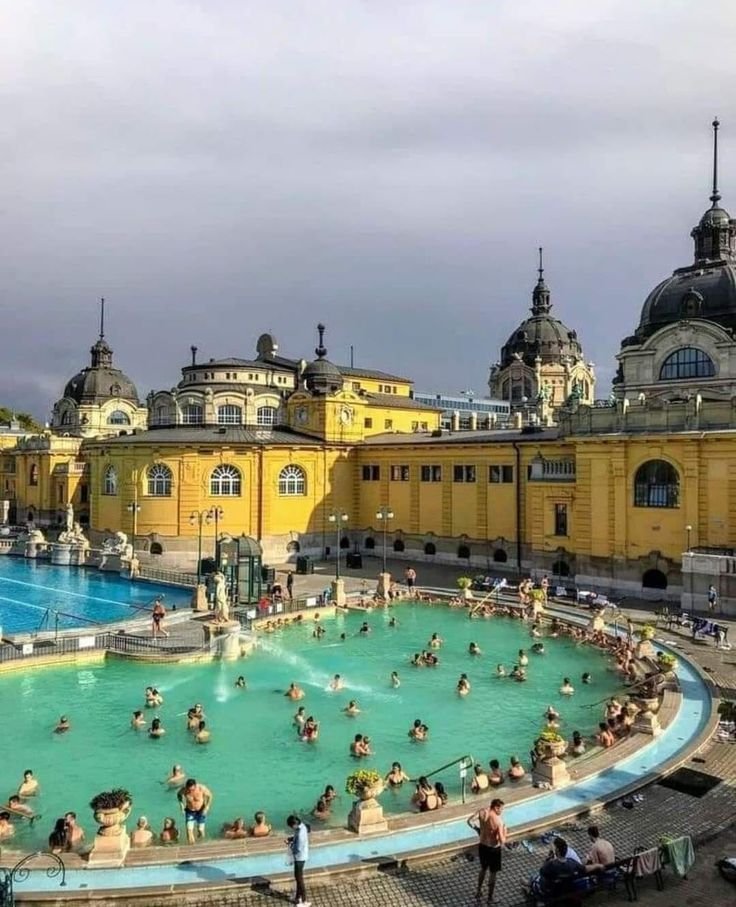 This may contain: many people are swimming in a large pool near some yellow buildings and green water fountains