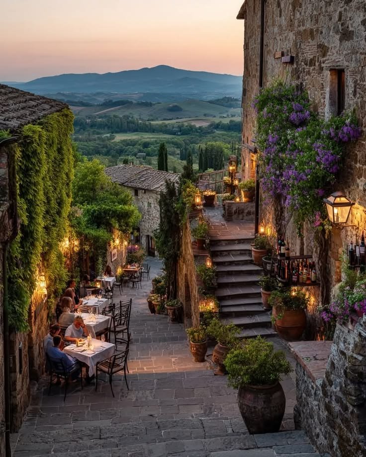 This may contain: an outdoor dining area with tables and chairs in the evening, surrounded by greenery