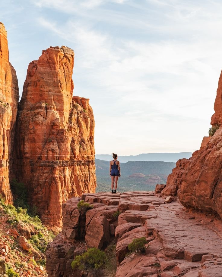 This may contain: a woman standing on the edge of a cliff in front of some red rock formations