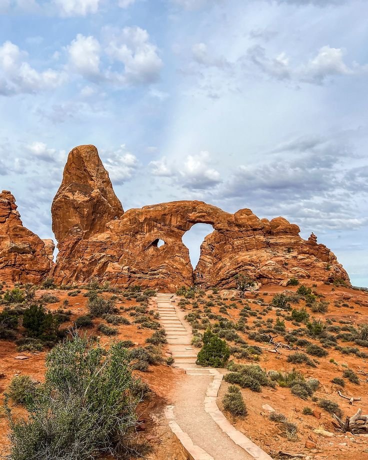 This may contain: an arch shaped rock formation in the desert with a path leading to it and trees on either side