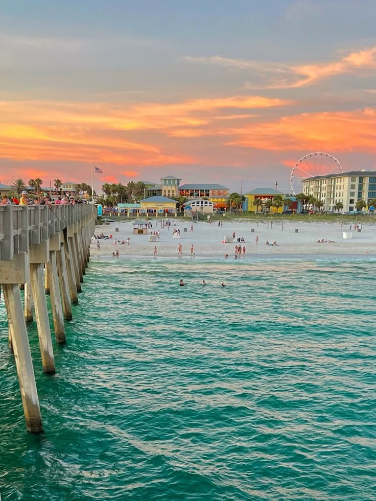 This may contain: people are swimming in the ocean near a pier at sunset or sunrise with ferris wheel in background