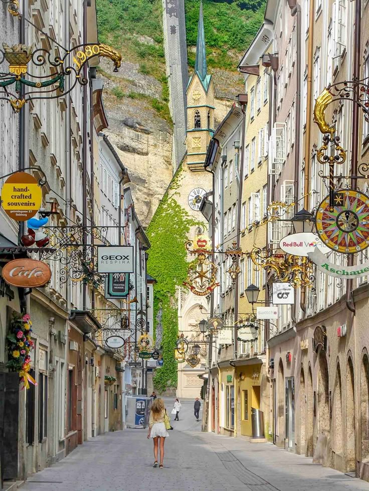 This may contain: a woman is walking down the street in an old european town with tall buildings and shops