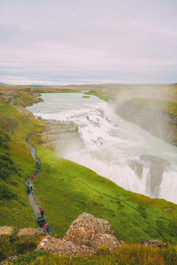 This may contain: people are walking along the edge of a cliff overlooking a waterfall and river with green grass on both sides