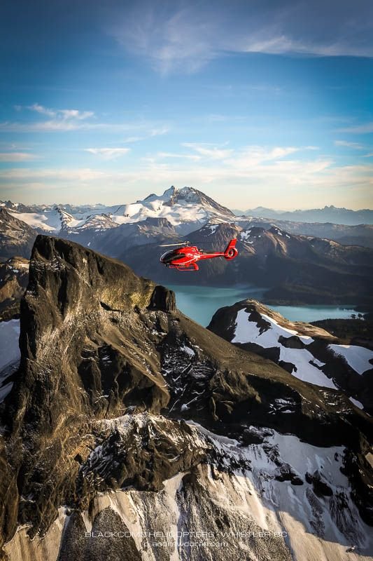 This may contain: a red helicopter flying over the top of a snow covered mountain with mountains in the background