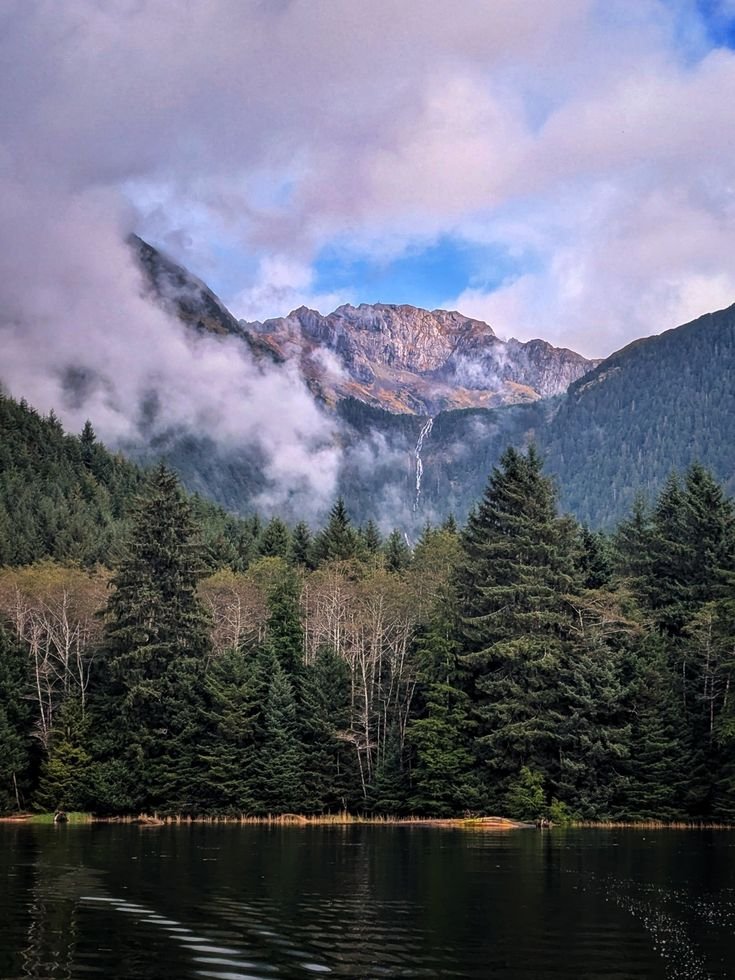 This may contain: the mountains are covered with clouds and trees in front of a body of water that is surrounded by evergreens