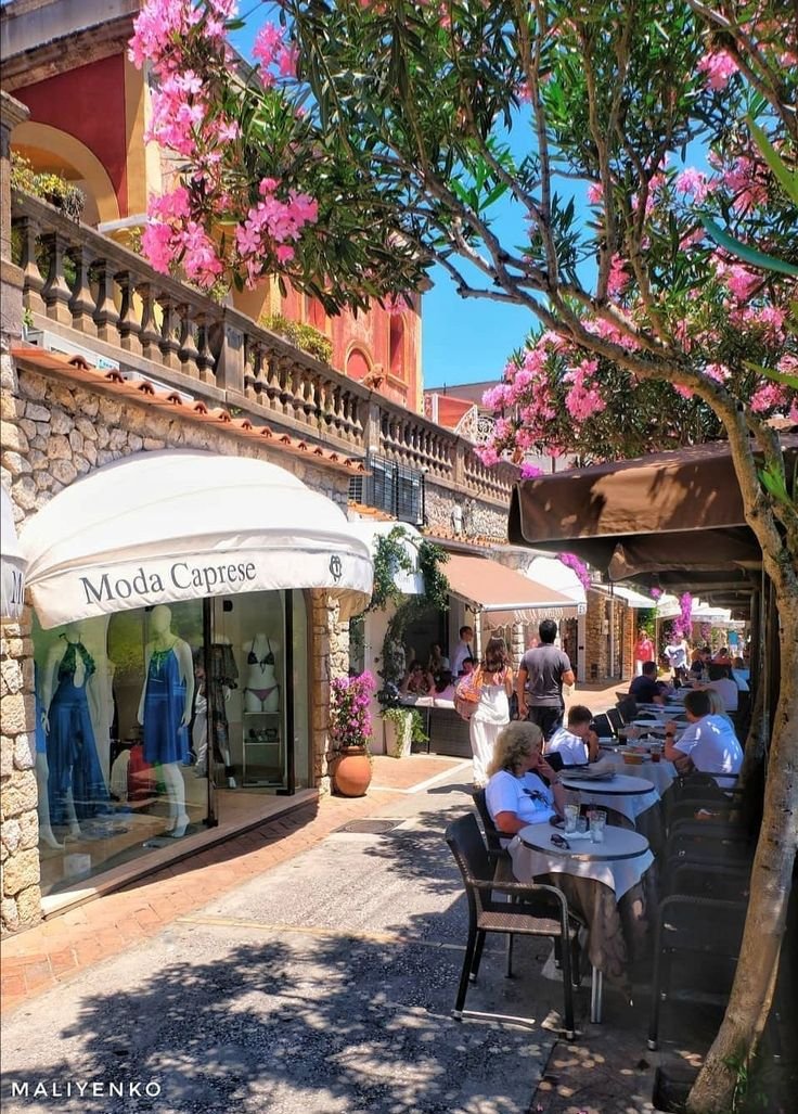 This may contain: people are sitting at tables under umbrellas in front of shops on the street with pink flowers