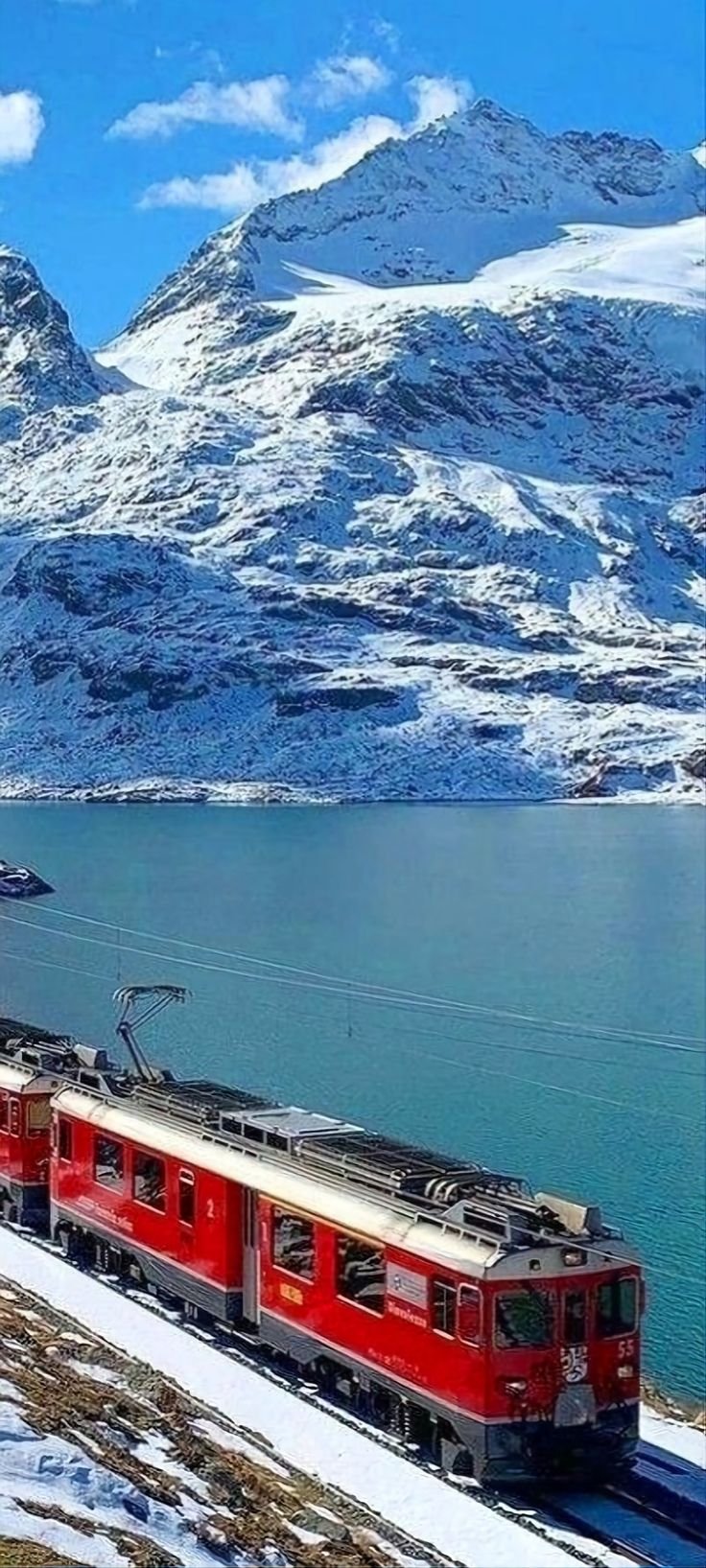 This may contain: a red train traveling down tracks next to snow covered mountains and water in the background