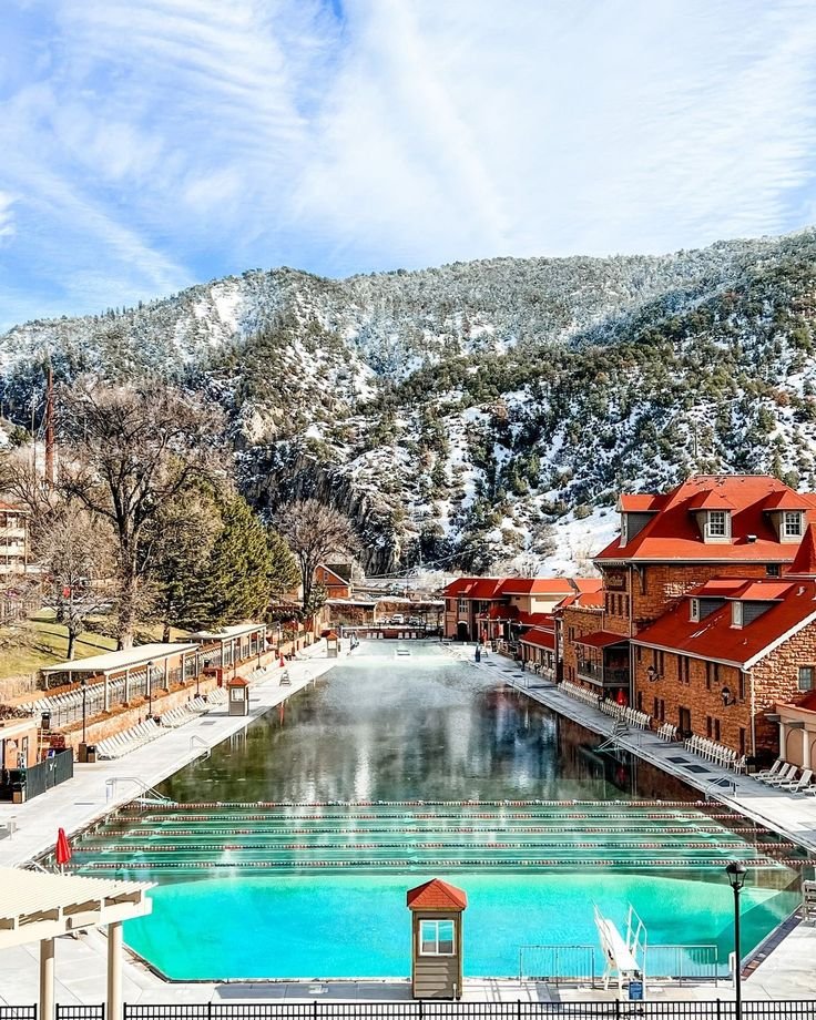 This may contain: an outdoor swimming pool surrounded by snow covered mountains and buildings in the distance with people walking around it