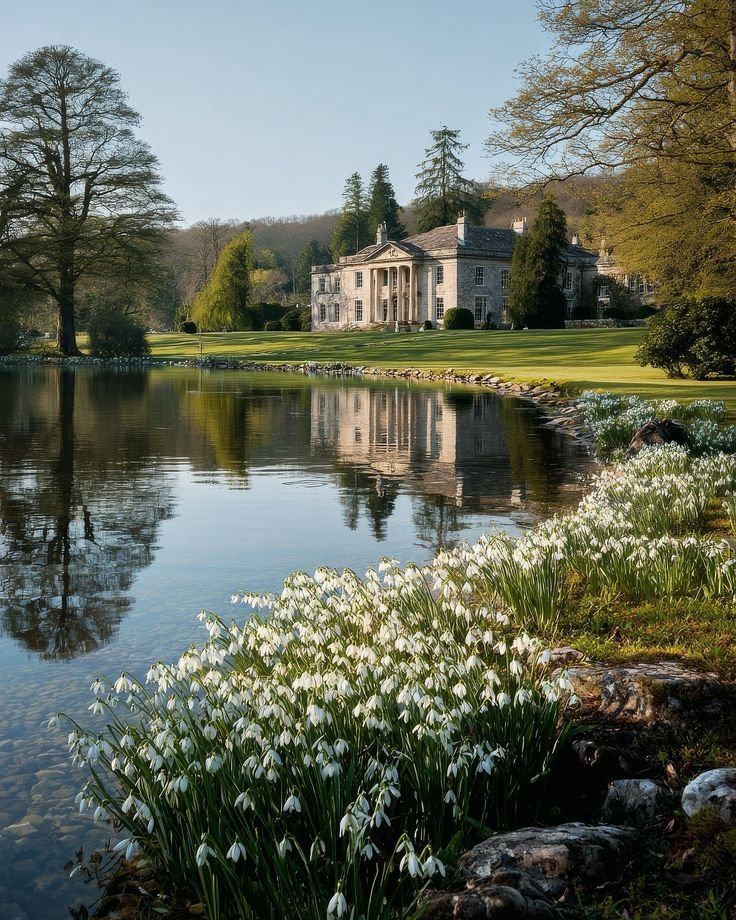 This may contain: a large house sitting on top of a lush green field next to a lake filled with flowers