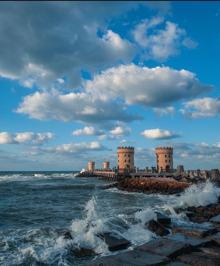This may contain: waves crashing against the rocks near an ocean shore with two buildings in the background and blue sky