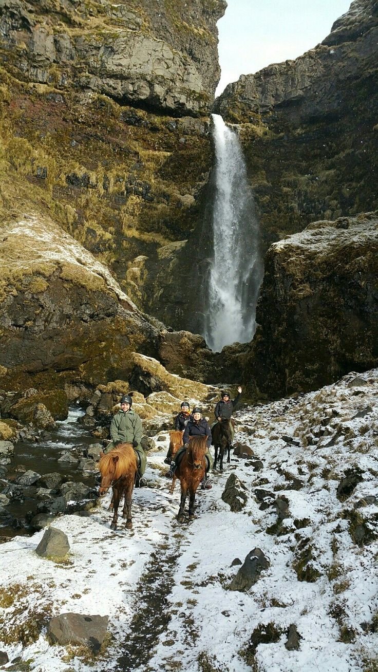 This may contain: several people riding horses in front of a large waterfall with snow on the ground and rocks