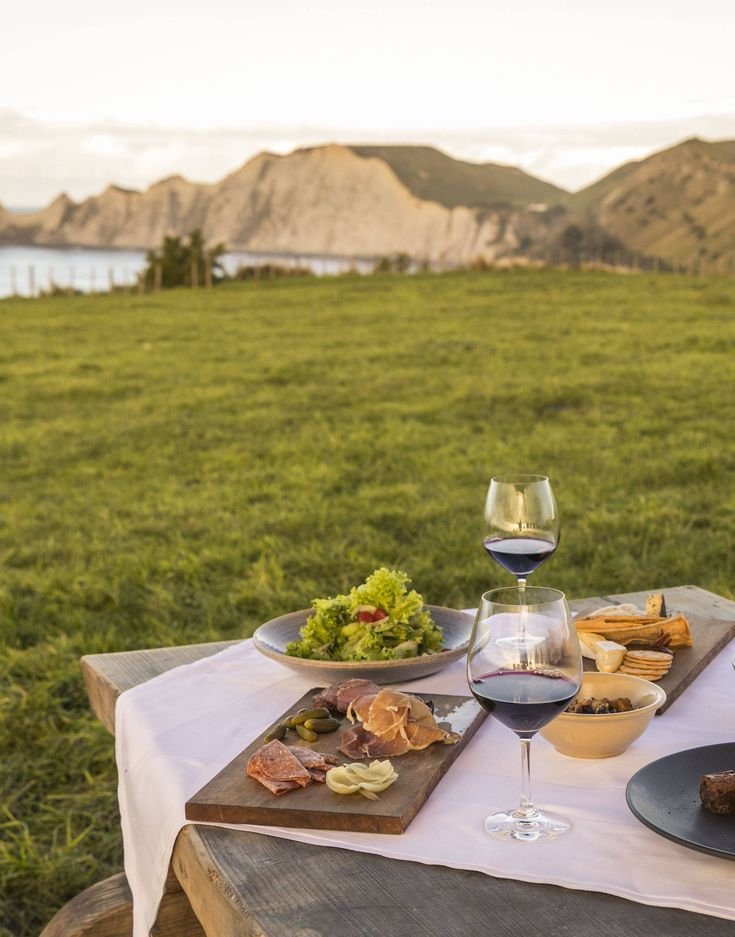 This may contain: two plates of food and wine on a picnic table in the grass with mountains in the background