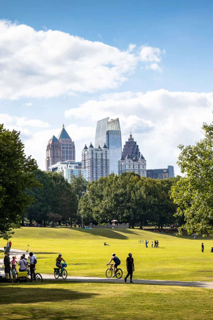 This may contain: people walking and riding bikes in a park with tall buildings behind them on a sunny day