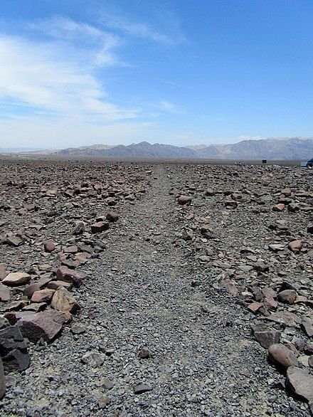 This may contain: a dirt road with rocks on the ground and mountains in the distance, under a blue sky