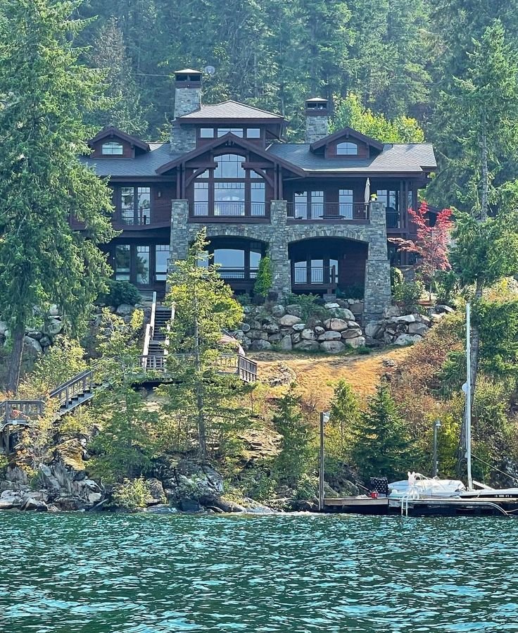 This may contain: a large house sitting on top of a lush green hillside next to a lake filled with lots of trees