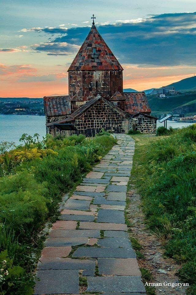 This may contain: an old church sits on the edge of a grassy hill next to a body of water