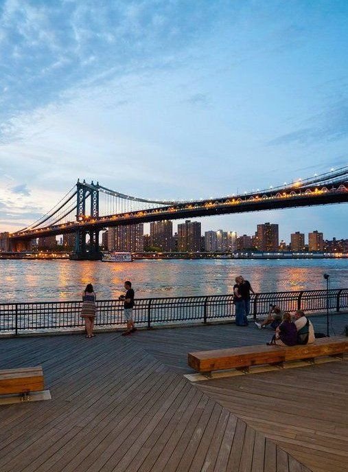 This may contain: people are sitting on benches near the water with a bridge in the background at dusk