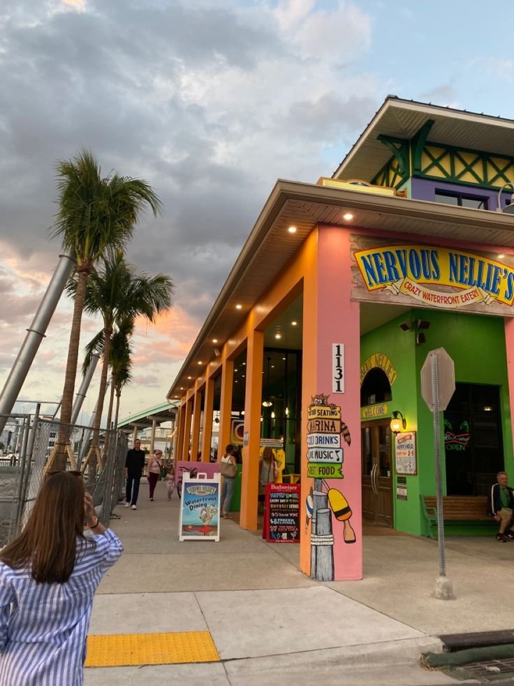 This may contain: a woman taking a photo of the front of a restaurant with palm trees in the background