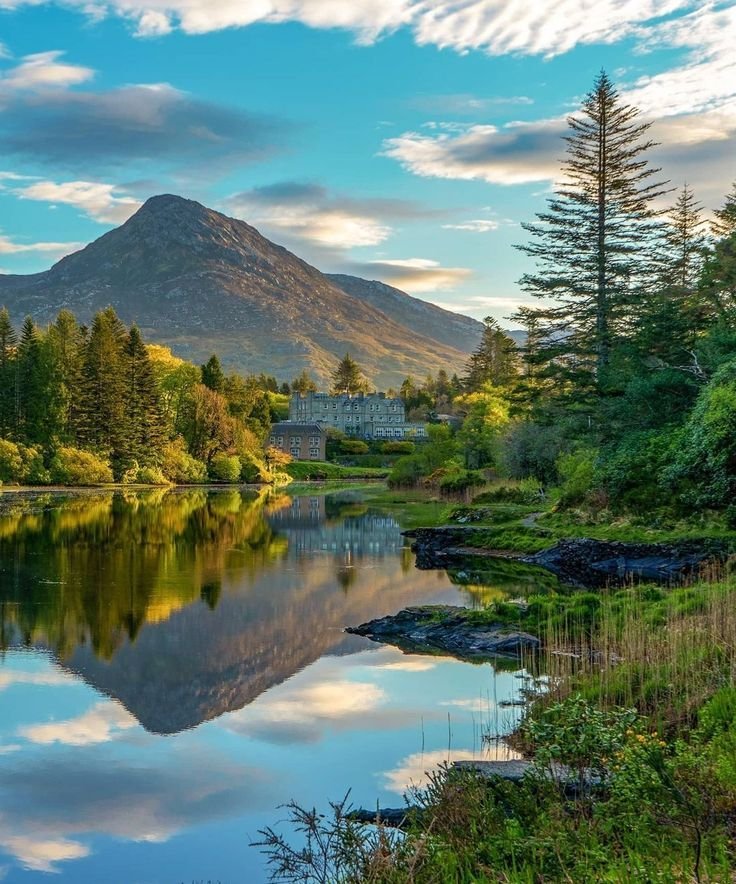This may contain: a lake surrounded by trees and mountains under a blue sky with clouds in the background