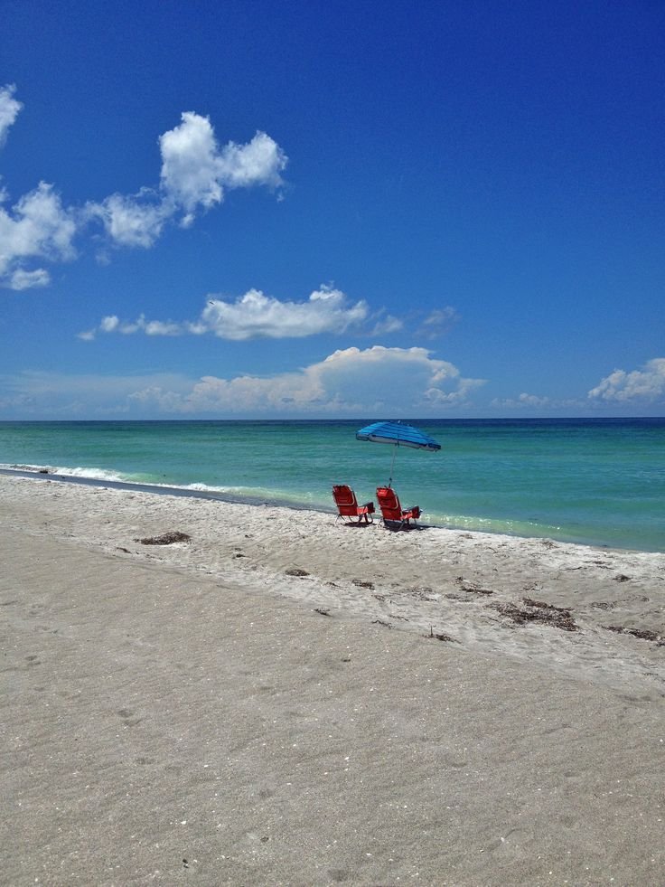 This may contain: two red lawn chairs sitting on top of a sandy beach next to the ocean under a blue umbrella