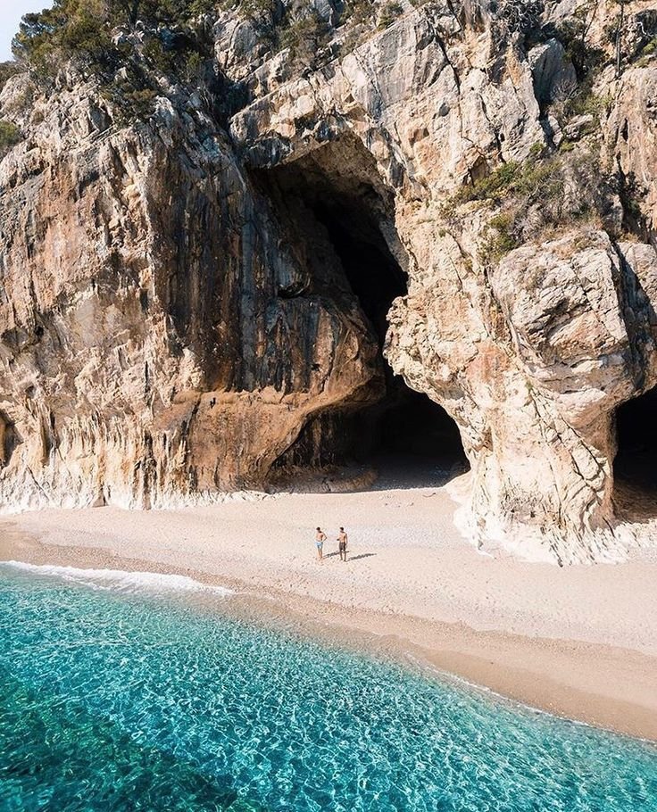 This may contain: two people standing on the beach in front of an ocean cave with clear blue water