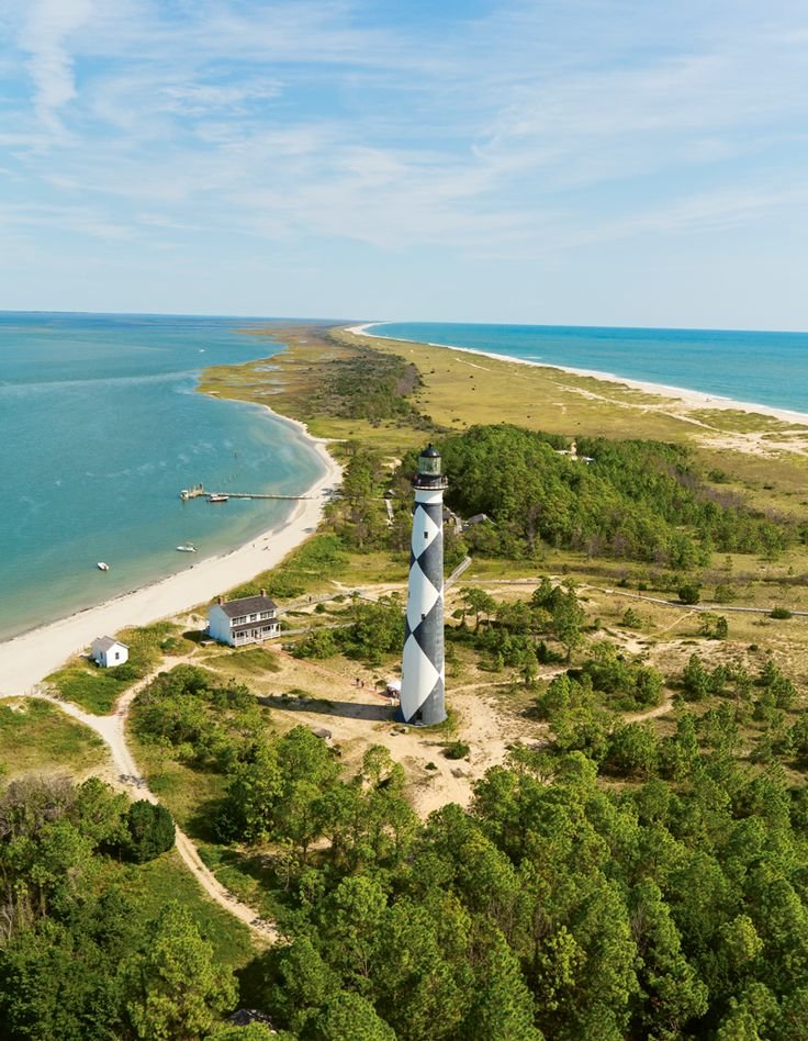 This may contain: an aerial view of a lighthouse surrounded by trees near the ocean and beach with boats