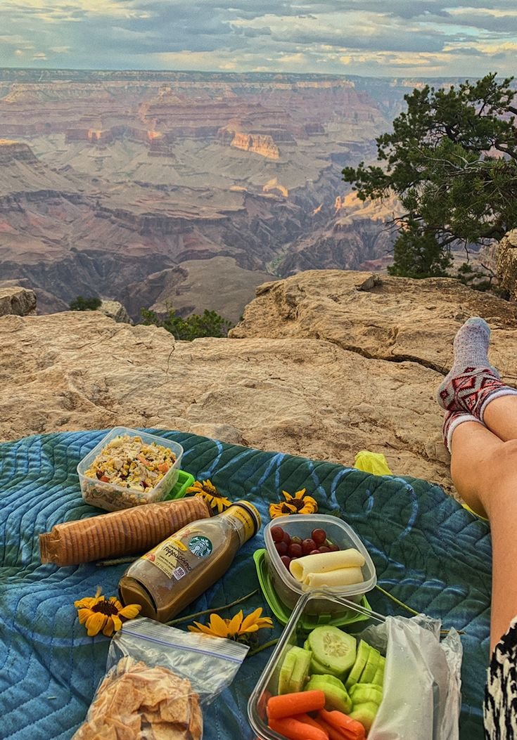 This may contain: a woman sitting on top of a blue blanket next to food and drinks at the edge of a cliff