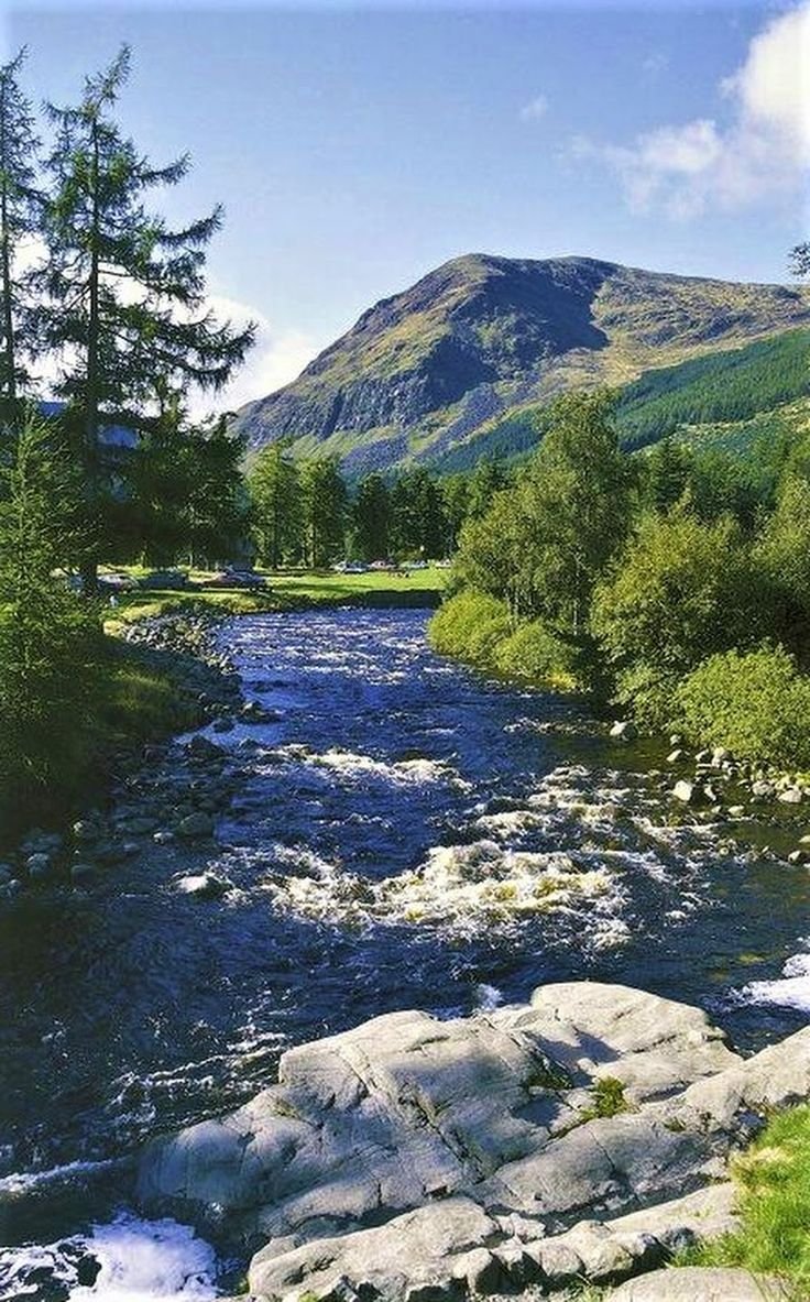 This may contain: a river running through a lush green forest covered hillside next to a tall mountain range