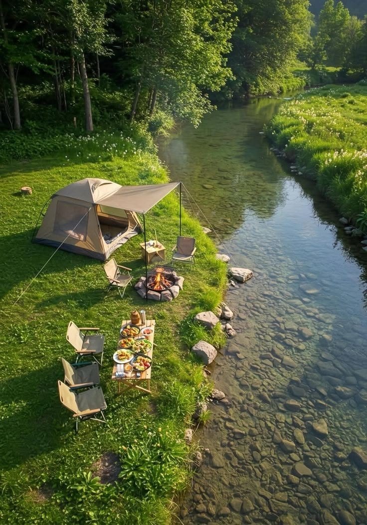This may contain: a tent set up next to a river with chairs and food on the grass near it