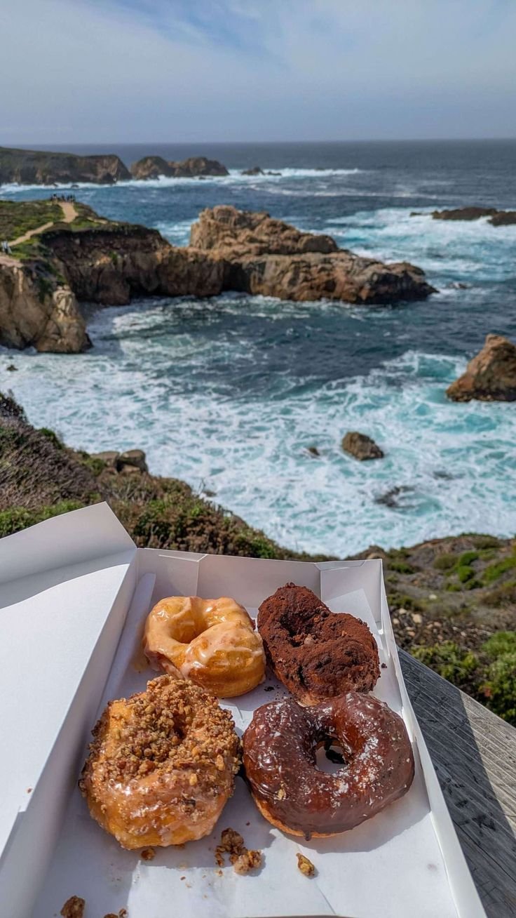 This may contain: three donuts are in a box on a table near the ocean and some rocks