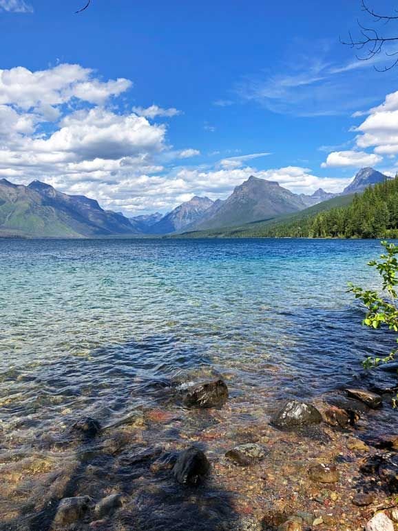 This may contain: a lake surrounded by mountains and rocks under a blue sky with clouds in the background
