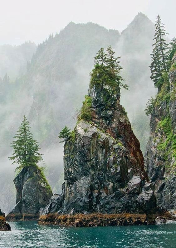 This may contain: an island in the middle of water surrounded by mountains and pine trees on a foggy day