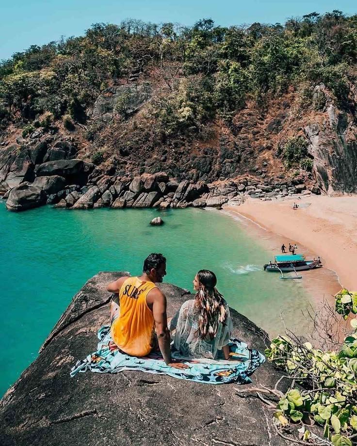 This may contain: two people sitting on top of a rock next to the ocean