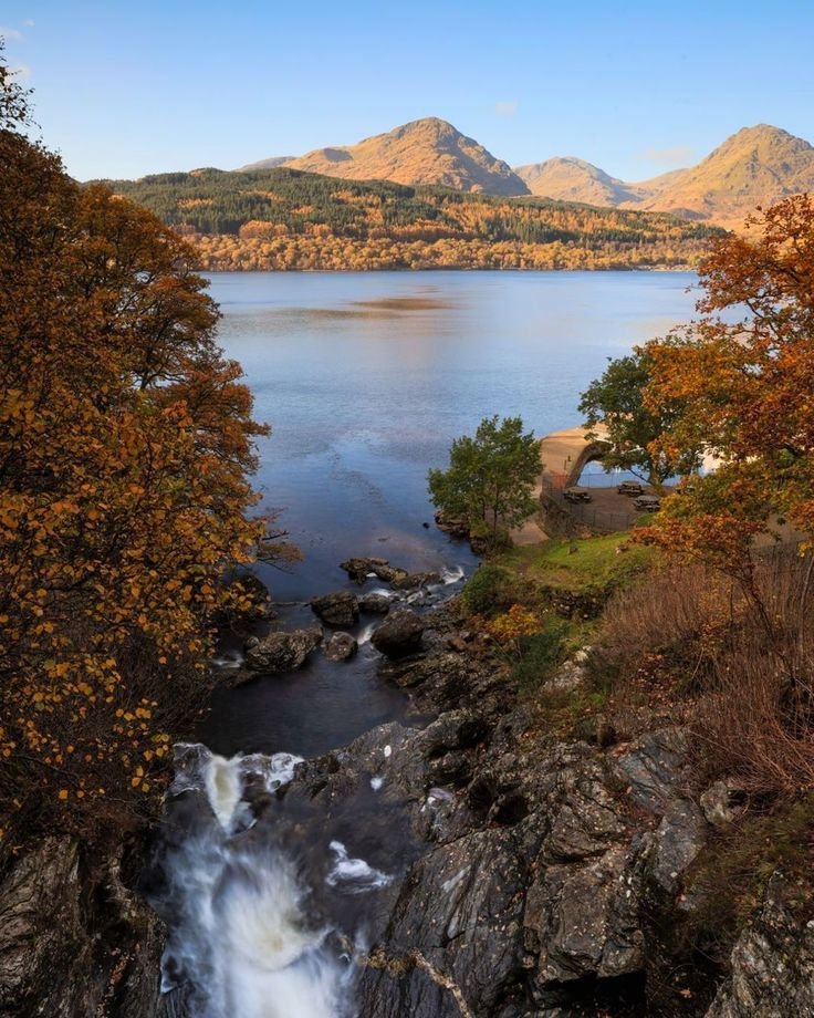 This may contain: a small waterfall in the middle of a lake surrounded by hills and trees with autumn foliage