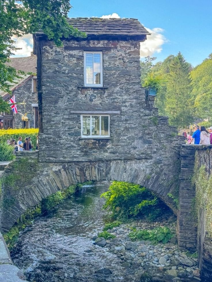 This may contain: people standing on the side of a stone bridge over a small river in front of a house