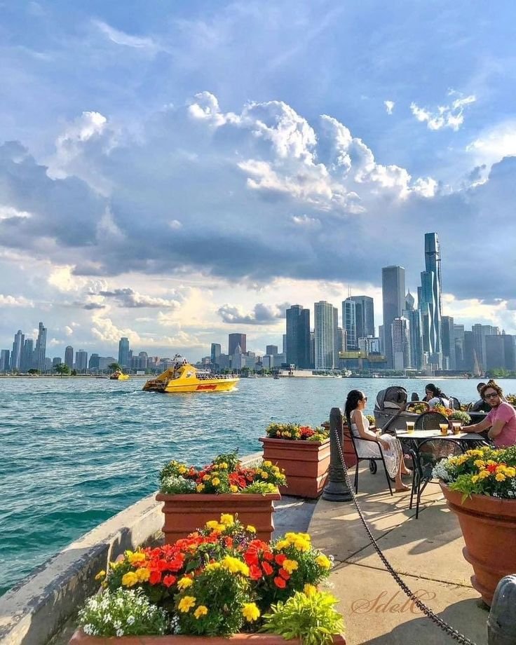 This may contain: people sitting at tables near the water in front of a cityscape with boats
