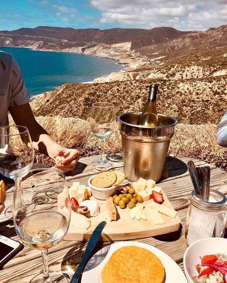 This may contain: two people sitting at a table with food and wine overlooking the ocean on a sunny day