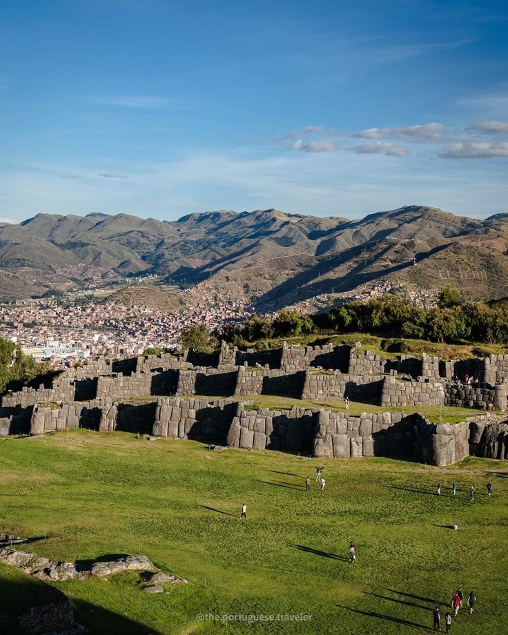 This may contain: people are walking around in the grass near an old stone structure with mountains in the background