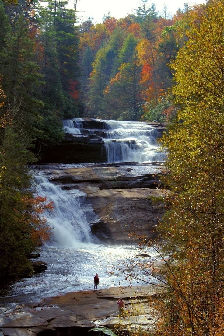 This may contain: two people are standing at the base of a waterfall with fall foliage around them and trees in the background