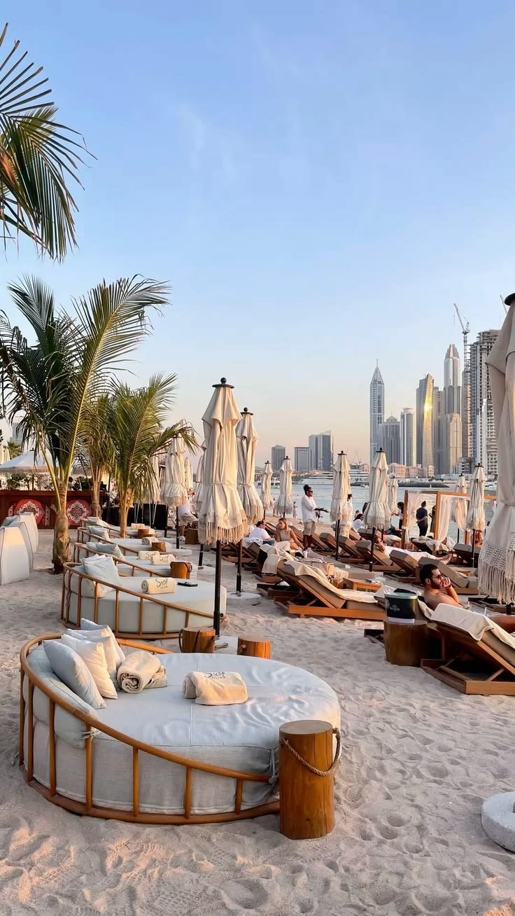 This may contain: many lounge chairs and umbrellas are on the sand near the water in front of a cityscape