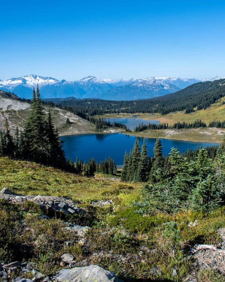 This may contain: a lake surrounded by mountains and trees on the side of a hill with snow capped mountains in the distance