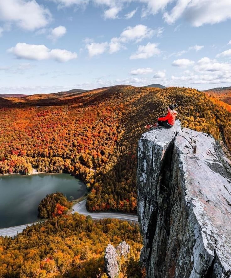 This may contain: a man sitting on top of a cliff next to a lake in the middle of autumn