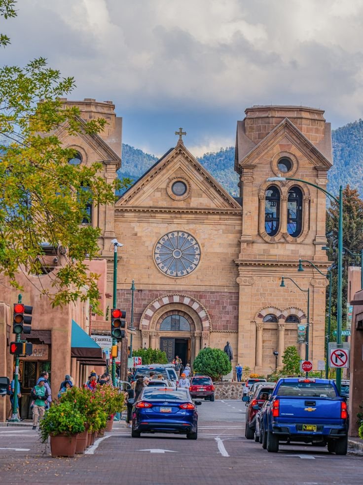 This may contain: cars parked in front of a church with a clock on it's face and mountains in the background