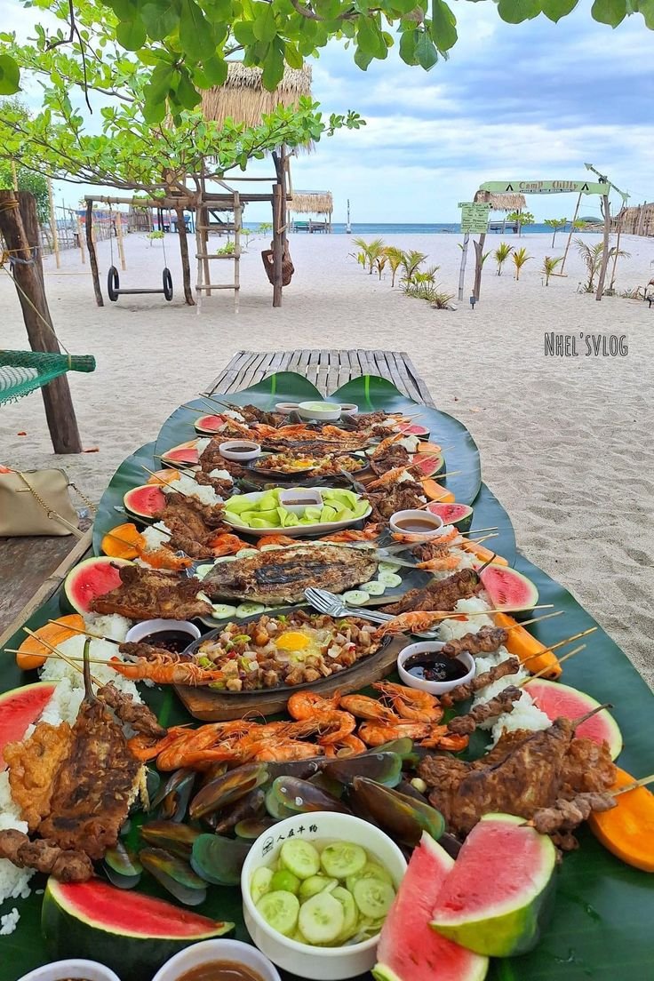 This may contain: a long table covered in lots of food on top of a beach next to the ocean