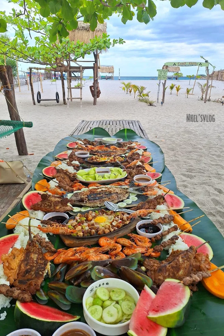 This may contain: a long table covered in lots of food on top of a beach next to the ocean