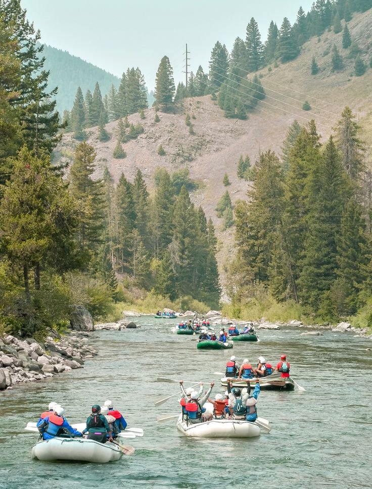 This may contain: several people are rafting down a river with mountains in the background and trees on either side