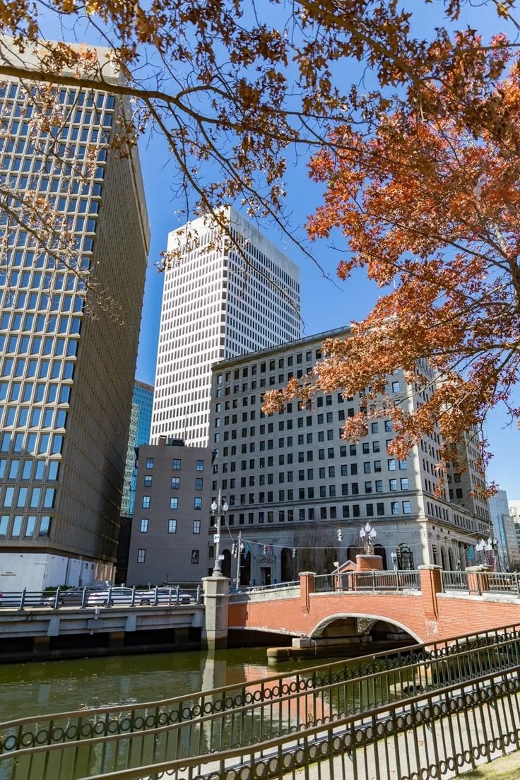This may contain: a bridge over a body of water in front of tall buildings and trees with leaves on them