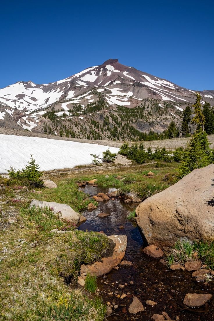 This may contain: a mountain with snow on it and rocks in the foreground, near a stream